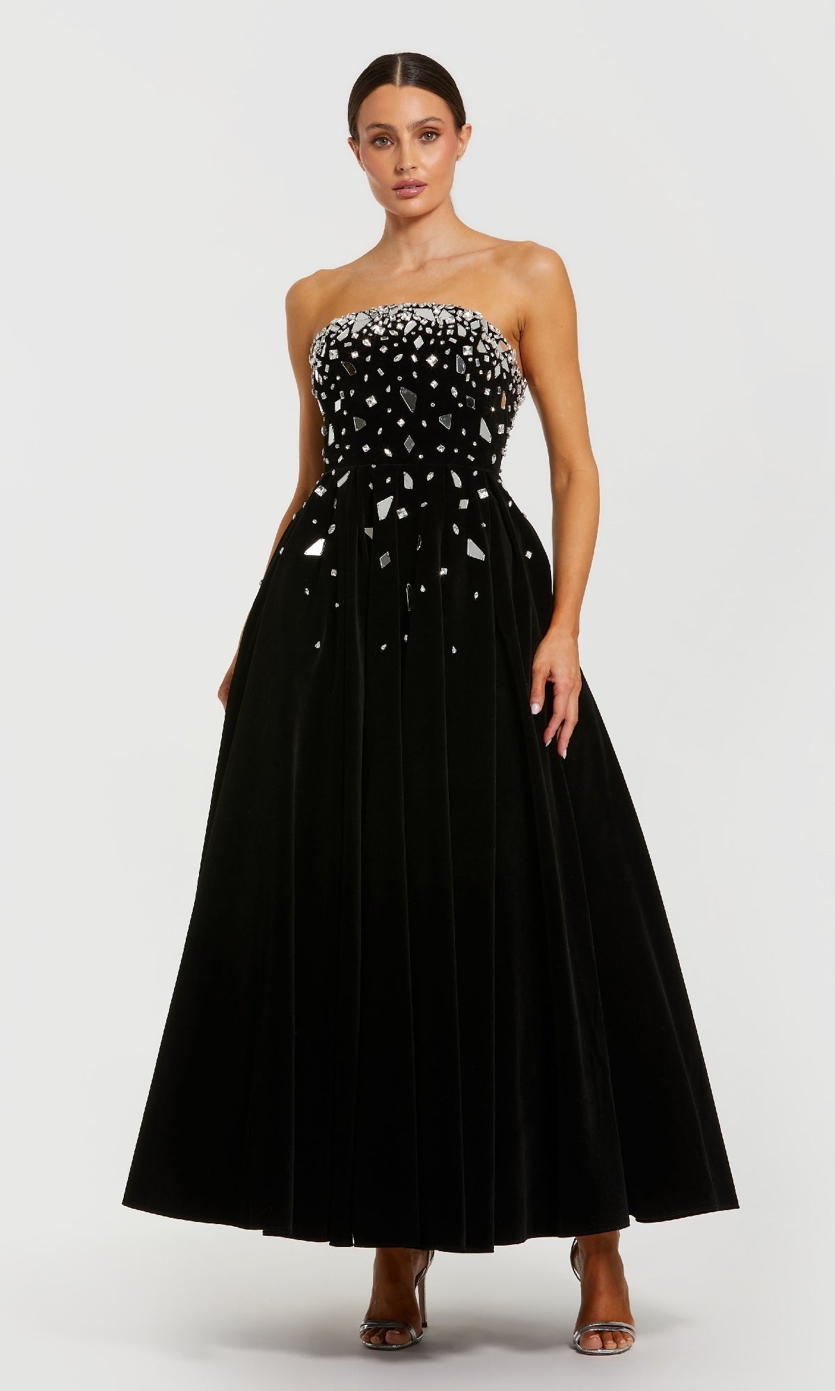 A woman wears the Mac Duggal 56197 long formal dress, a strapless black velvet gown with a rhinestone-embellished bodice featuring silver geometric accents and a pleated floor-length skirt, paired with silver open-toe heels.